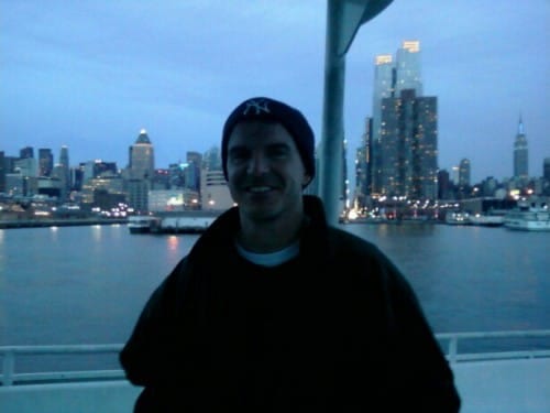 A cute guy in a NY Yankees knit cap at dusk with several lighted buildings behind him