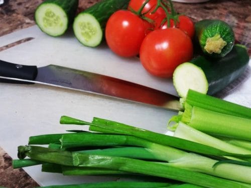 A white cutting board with sliced cucumbers, tomatoes, zucchini, and green onions