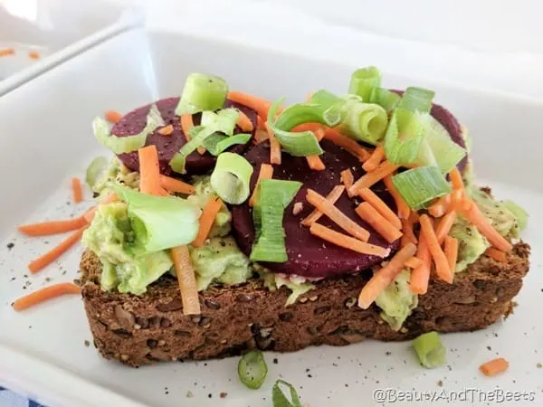 A slice of bread with mashed avocado, grated carrots, sliced red beets and green onions on a square plate