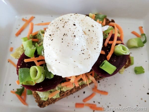 A large poached egg on top of green onions, sliced red beets, grated carrots and mashed avocado on a white square plate