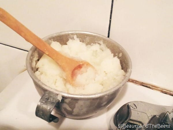 a silver cup filled with sugar scrub and a wooden spoon on a white counter with a white tile wall