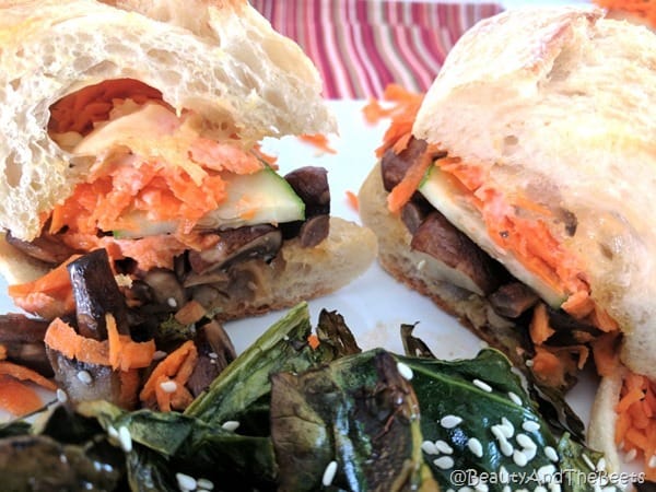 a colorful sandwich with shredded carrots and cucumbetrs on crusty bread with a serving a roasted greens in the foreground on a red striped placemat