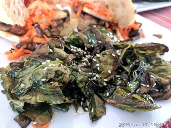 a serving of roasted gai lan greens with white sesame seeds and a colorful sandwich in the background on a white plate