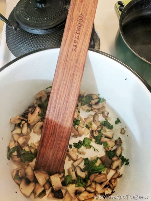 a white stock pot with sauteed mushrooms and mustard greens with a wooden Earlywood spatula with a black cast iron tea pot in the background on a white stove