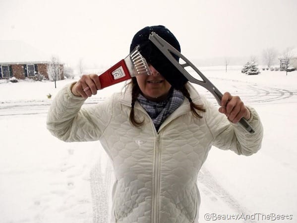 the author wearing a white jacket and black knit cap with snow removal brush and scraper covering her eyes with a snowy background