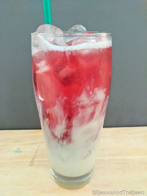 a tall glass of an mbre drink with a light green milky bottom and pinkish red tea on top over ice on a dark gray background