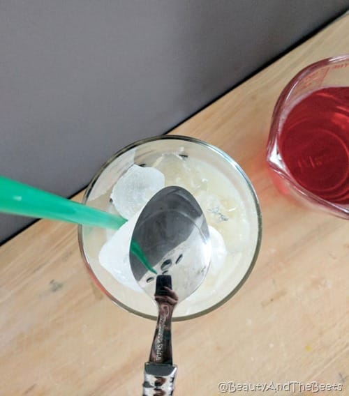 a glass with a green straw and ice with the backside of a spoon over top next to a glass of red tea