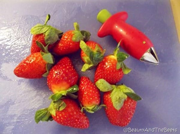 a small bunch of strawberries next to a strawberry huller gadget on a blue cutting board