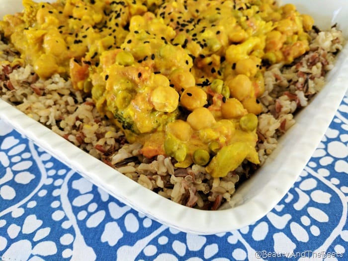 a white dish filled with a yellow curried vegetables and chickpeas with a sprinkling of black sesame seeds and brown grain rice in a white dish on a blue and white patterened placemat