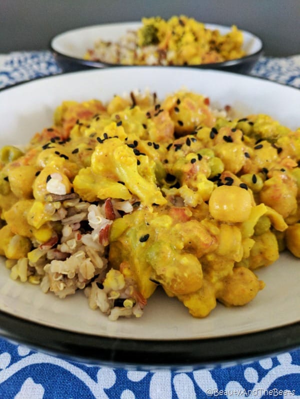 a white bowl with a black trim filled with yellow curried vegetables mostly caulifower with a same bowl in the background on a white and blue patterned placemat