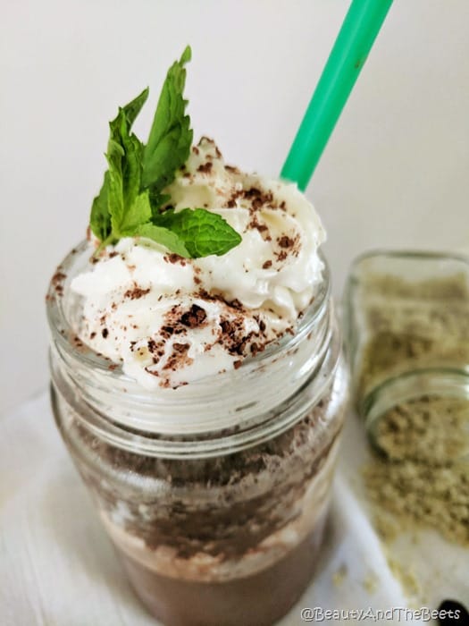 a mason jar with chocolate drink and whipped cream with a mint leaf and cocoa powder with a green straw on a white tablecloth