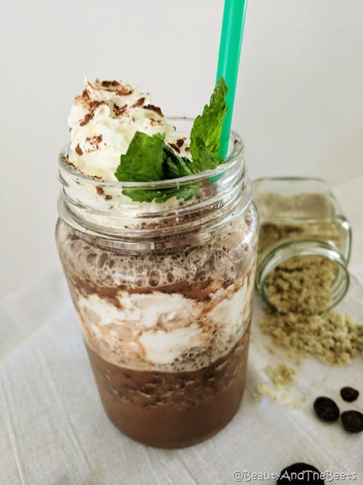 a mason jar with chocolate drink and whipped cream with a green straw on a white tablecloth with a tipped jar of mint sugar next to the drink