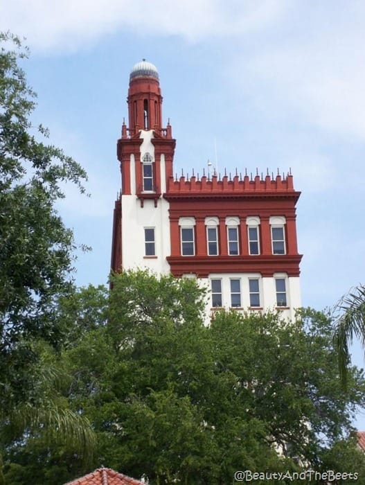 a view of a brown and white spanish style building peering out over the trees against a bright blue sky