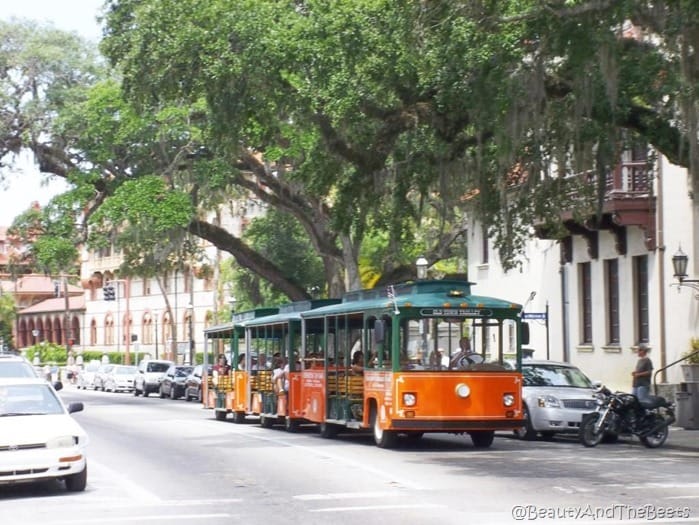 an orange trolley with a green roof on a street in St augustine with spanish architecture in the background