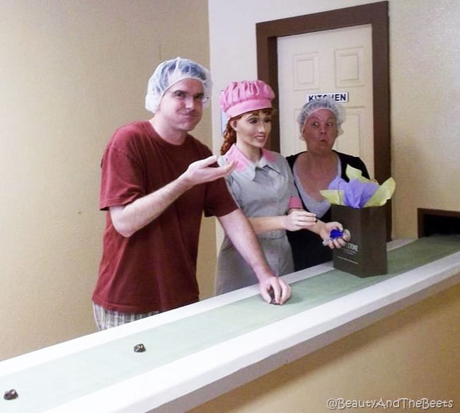 Mr Beet in a red shirt with a hairnet and the author in a hairnet caught eating a chocolate with puffy cheeks standing on either side of a Lucille Ball statue on a chocolate belt line