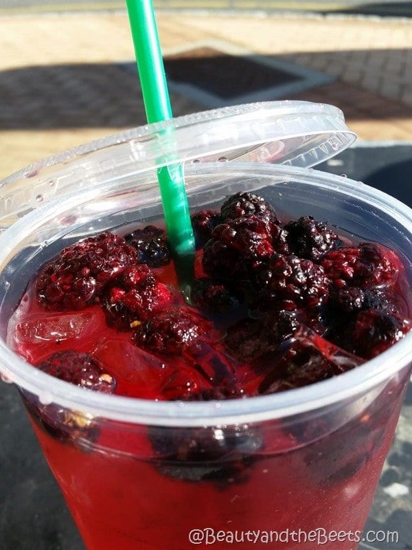 A large plastic cup filled with a red refresher drink and a pile of blackberries on top with an open lid and a green straw