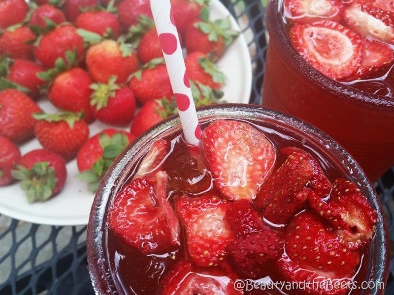 A glass filled with a red iced drink and strawberries in the drink. A red and white polka dot straw is in the glass. In the background is a plate of fresh red strawberries. Strawberry Acai Refresher recipe