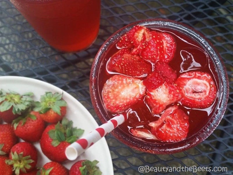 An overhead shot of a red iced drink with a red and white polka dot straw on an outdoor table with a white plate of fresh red strawberries