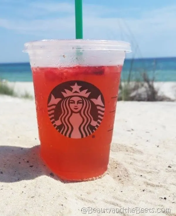 A light red iced drink in a Starbucks cup with a green straw on a white sandy beach with the blue green water in the background. Strawberry Acai Referesher recipe