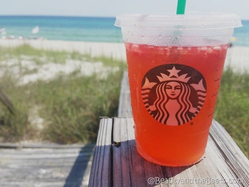 The Starbucks copycat Straberry Acai Refresher recipe  - A red drink with ice in a Starbucks cup with a green straw on the wood walkway heading towards the beach. The beach grass and white sand is in the background