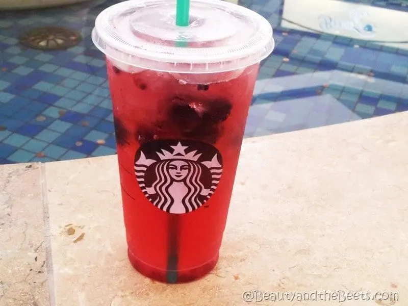A large plastic Starbucks cup of the red refresher drink in front of a blue tiled fountain with pennies in the water