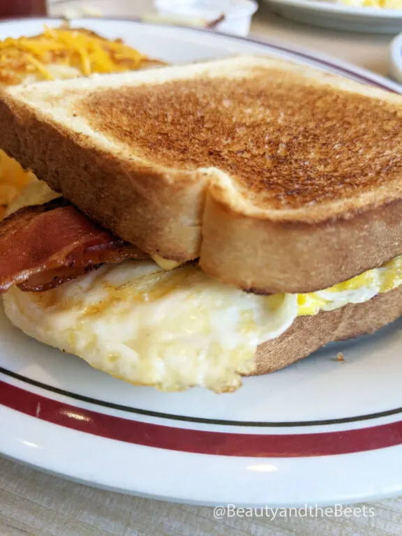 a large piece of toasted bread with bacon, egg, and cheese on a white plate with a maroon and green striped border