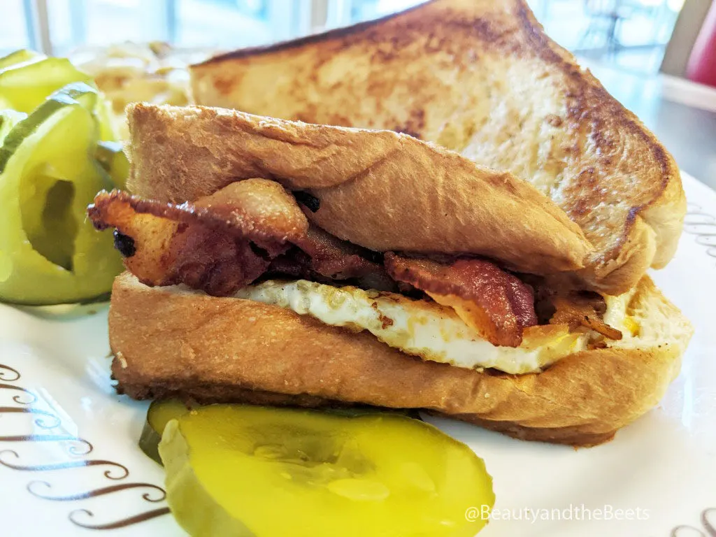 Thick bread sandwich with bacon, egg, and cheese on a white plate with a large slice of pickle in front of the sandwich and a small pile of pickles behind the sandwich