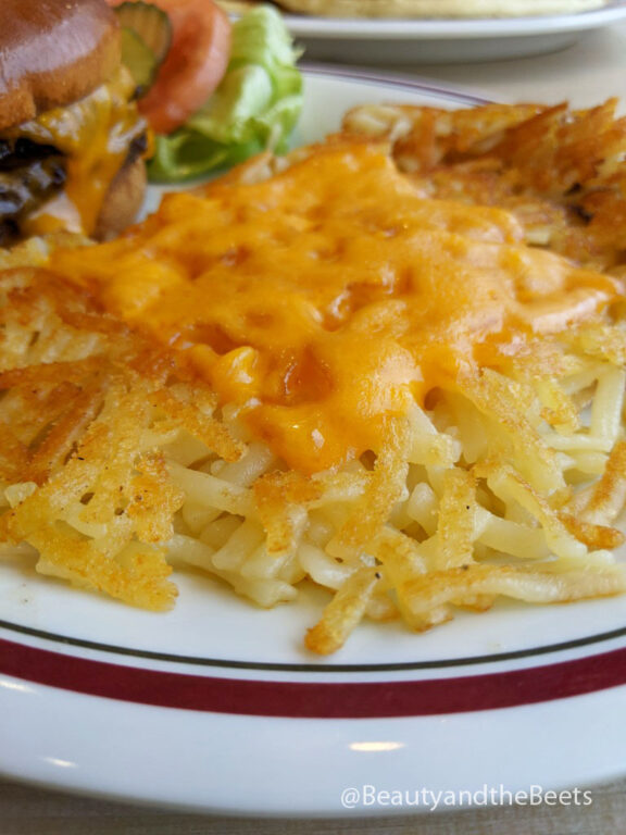 an up-close photo of hashbrowns topped with a slice of melted American cheese on a white plate with a maroon and green border