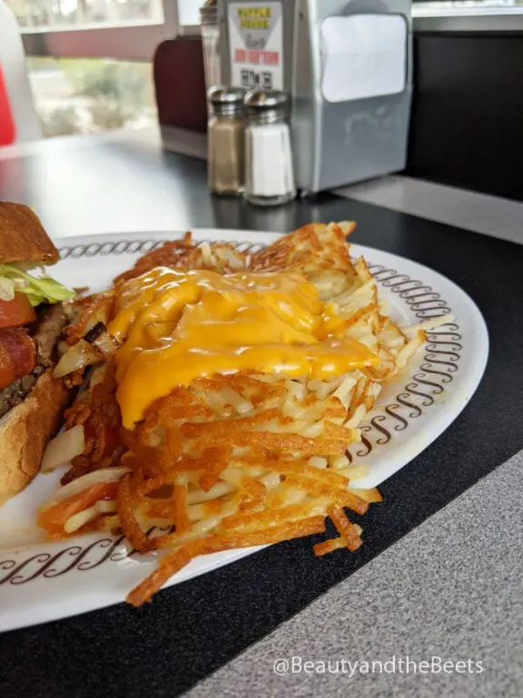 a column of crispy hashbrowns with a slice of melted American cheese at the edge of a white plate with a brown squiggly border in front of a metal napkin dispenser and a set of glass salt and pepper shakers