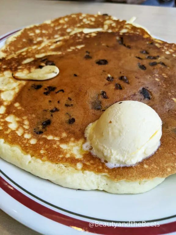 a white plate with a maroon and green striped border holding a large golden pancake sprinkled with chocolate chips and large dollop of butter