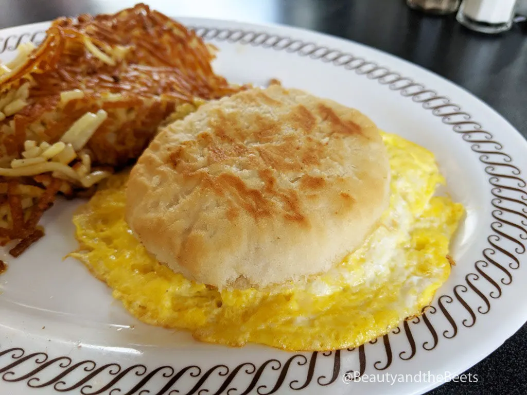 a golden biscuit sandwich with a large overflowing cooked egg next to a pile of crispy hashbrowns on a white plate with a brown squiggly border