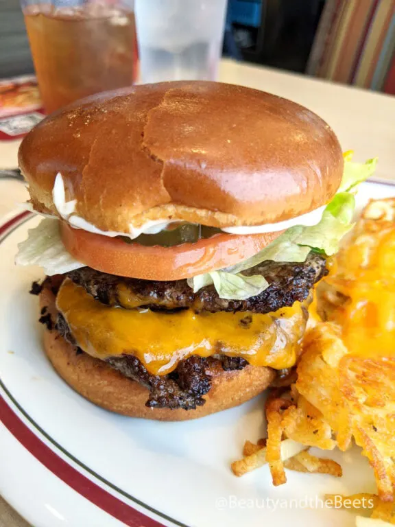 a big double cheeseburger with lettuce, tomato, pickles, and yellow cheese beside some crispy hashbrowns and melted cheese on top on a white plate with a maroon and green striped plate