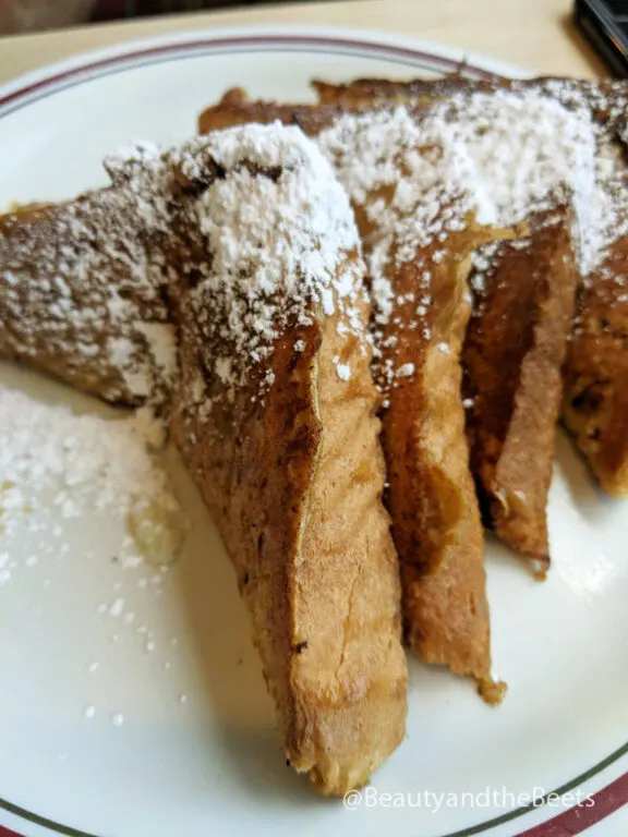 Four slices of thick French toast topped with a sprinkling of powdered sugar on a white plate with a maroon and green stripe border