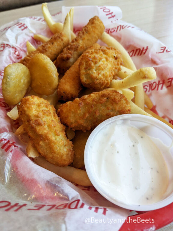 a pile of golden fried pickles, mozzarella sticks, and jalapeno poppers on a sheet of Huddle House imprinted paper with a large plastic ramekin of ranch sauce 