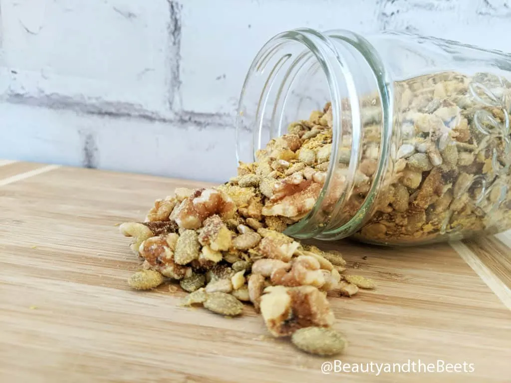 Salad Topper Recipe A small mason jar tipped over on its side with walnuts, pepitas, sunflower seeds, and nutritional yeast spilling out of it in front of a white brick background