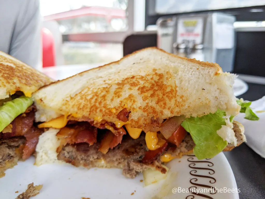 A  half burger with bacon, yellow cheese, green lettuce, and tomato on white toasted bread facing the camera on a white plate with a brown squiggly border