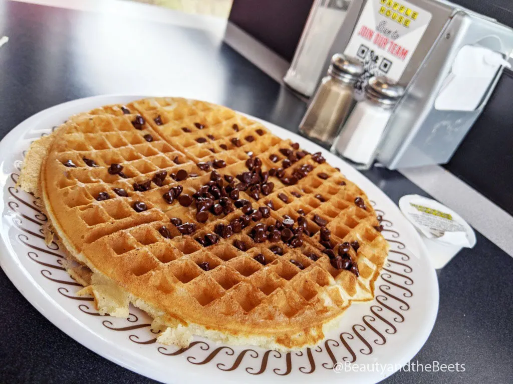 a large golden waffle with a sprinkling of chocolate chips on a white plate with a brown squiggle pattern border in front of a silver napkin dispenser with a pair of glass salt and pepper shakers