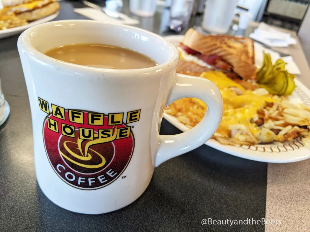 a white coffee cup with the Waffle House lettering logo and the word coffee on a dark gray table with a plate of cheesy hashbrowns in the background