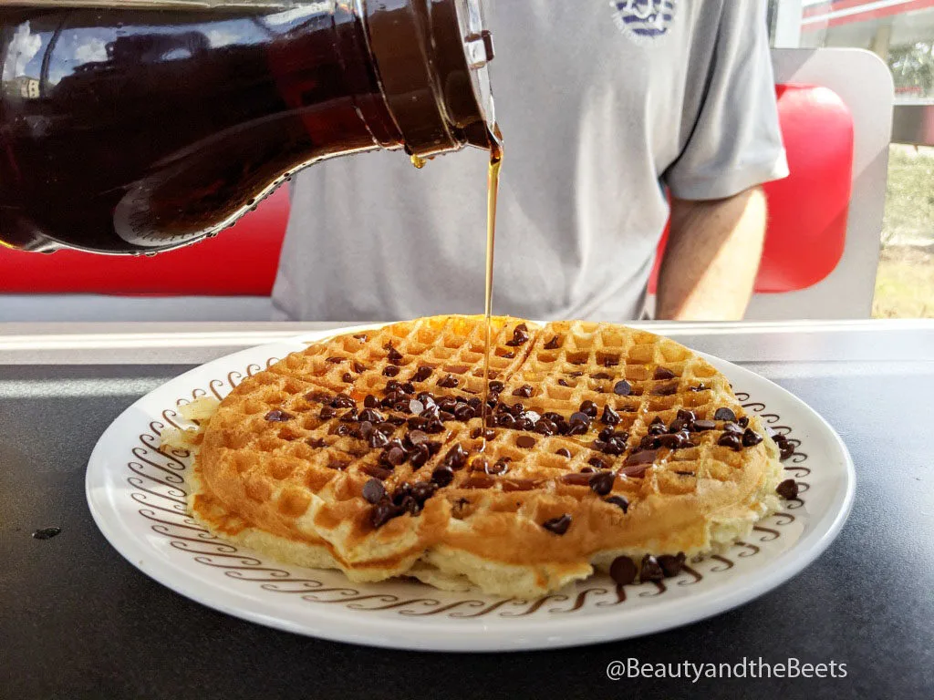 a man in a gray tee shirt in the background pouring syrup on a large white plate with a golden waffle on a white plate with a brown squiggle border on a gray top table with a red booth cushion behind the man