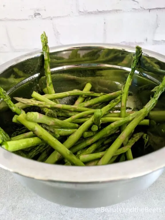 a silver mixing bowl with raw asparagus spears with white brick background