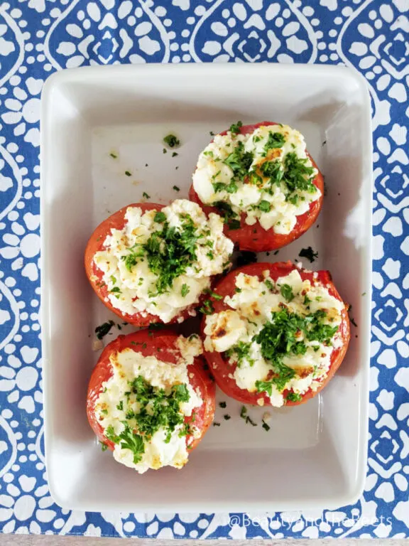 An overhead shot of four baked tomatoes with goat cheese and green parsley on a white square dish on a blue and white patterned countertop