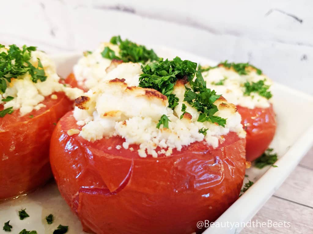A close up of a red blistered tomato with goat cheese and bright green parsley on a white rectangular dish on a light wooden counter top