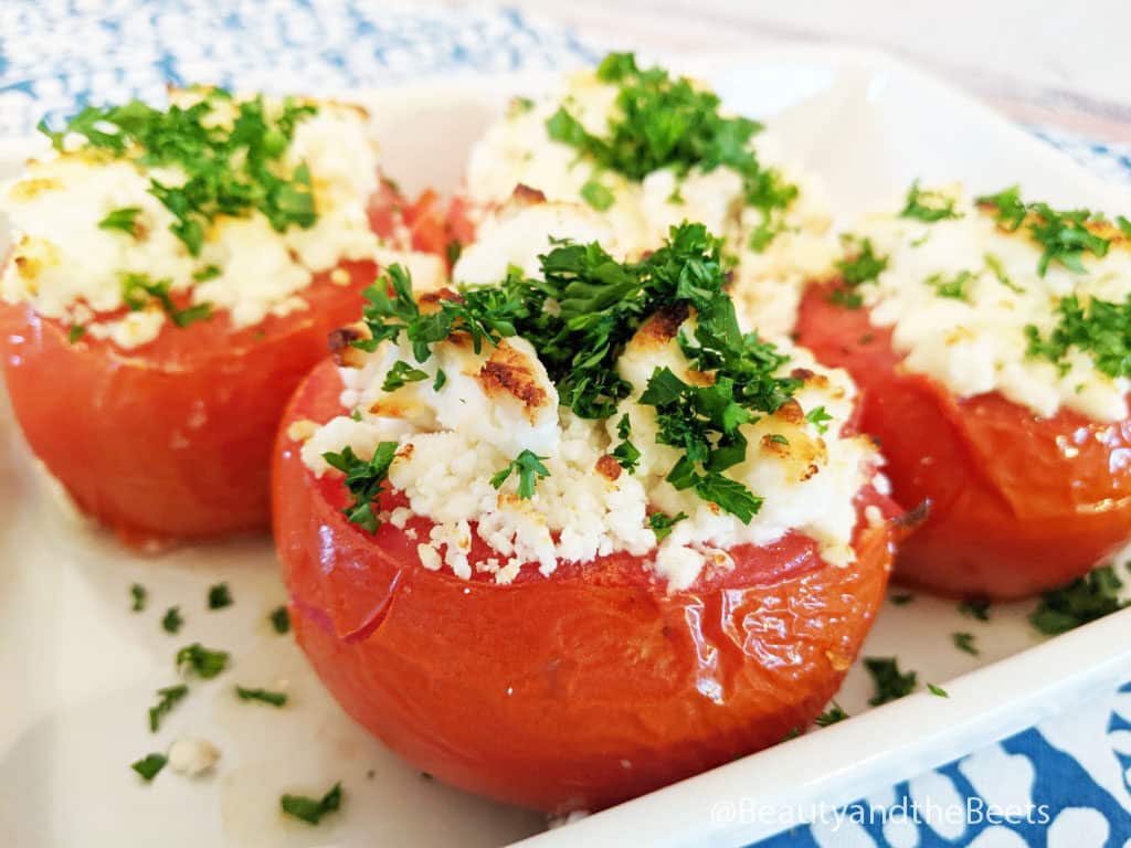 Baked tomatoes with goat cheese topped with parsley on  white plate placed on top of a blue and white patterned countertop