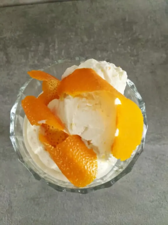 an overhead shot of a glass on a gray stone tabletop with a scoop of vanilla ice cream and bright orange curled peel