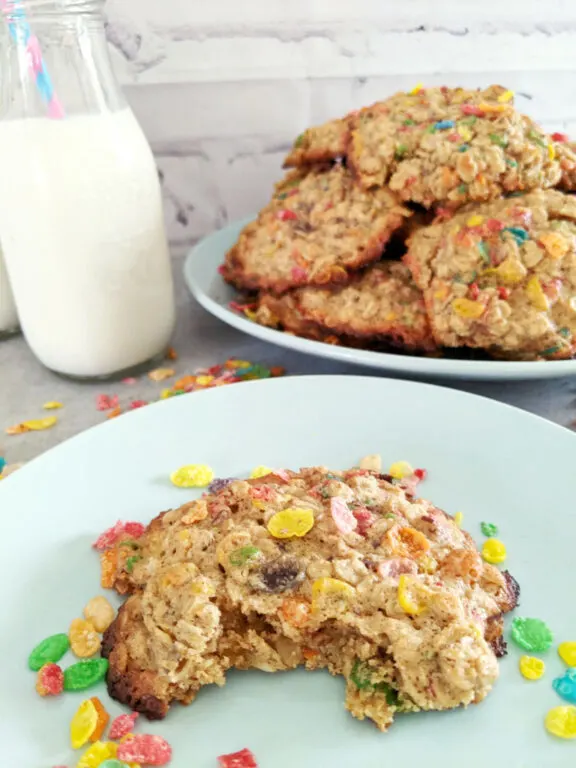 A colorful fruity pebbles breakfast cookie with a bite missing on a light blue plate with a sprinkling of fruity pebbles cereal in front of a plate of cookies and two mini jugs of white milk