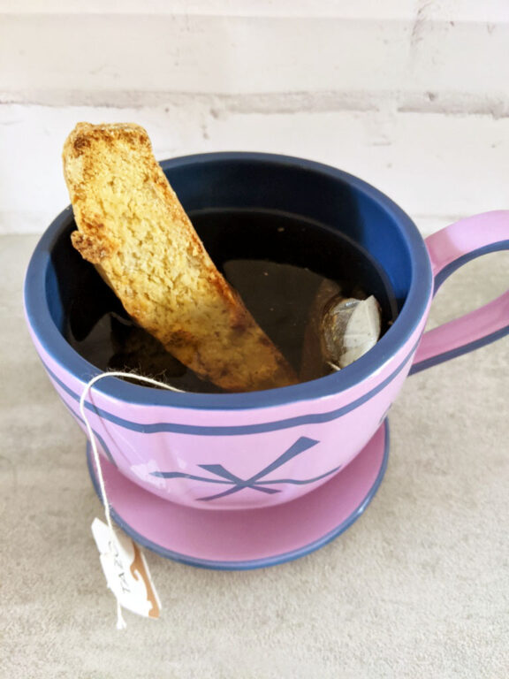 A biscotti cookie resting a cup of tea in a purple tea cup on a gray stone countertop